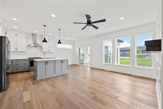 kitchen featuring appliances with stainless steel finishes, white cabinetry, an island with sink, hanging light fixtures, and wall chimney range hood