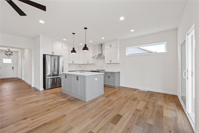 kitchen featuring stainless steel refrigerator, an island with sink, white cabinets, hanging light fixtures, and wall chimney exhaust hood