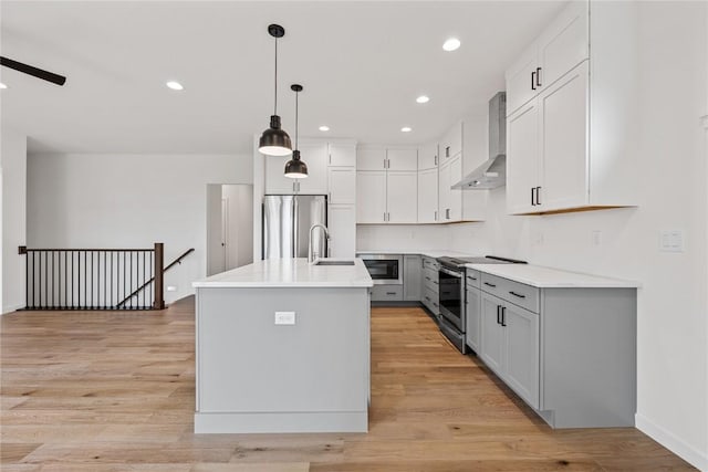 kitchen with sink, hanging light fixtures, appliances with stainless steel finishes, a kitchen island with sink, and wall chimney range hood