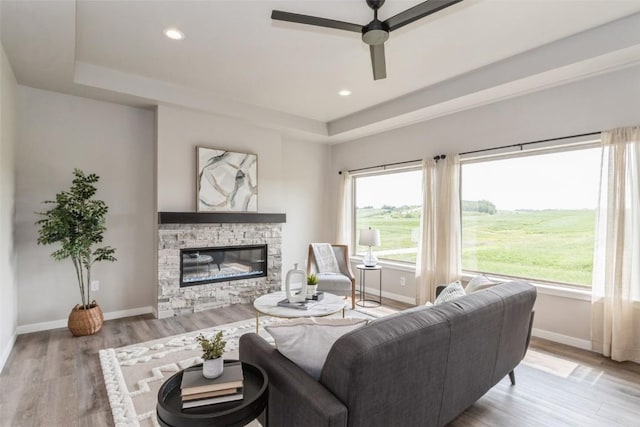 living room with ceiling fan, a tray ceiling, light hardwood / wood-style floors, and a stone fireplace