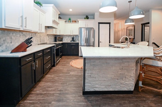 kitchen featuring appliances with stainless steel finishes, a kitchen island with sink, white cabinets, and decorative light fixtures