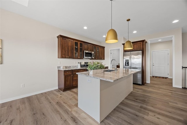 kitchen featuring appliances with stainless steel finishes, hanging light fixtures, light stone counters, an island with sink, and light wood-type flooring