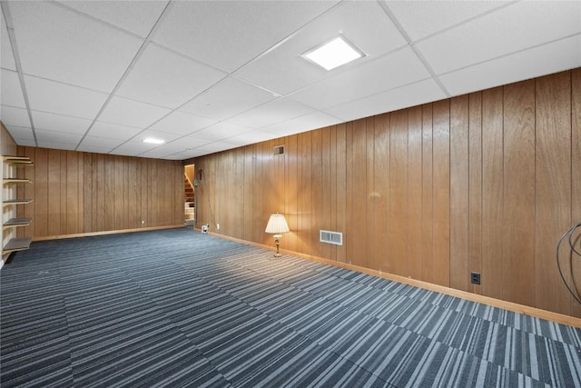 basement featuring a paneled ceiling, wooden walls, and dark colored carpet