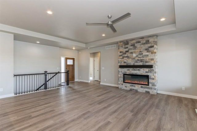 unfurnished living room featuring ceiling fan, a tray ceiling, a stone fireplace, and light hardwood / wood-style flooring