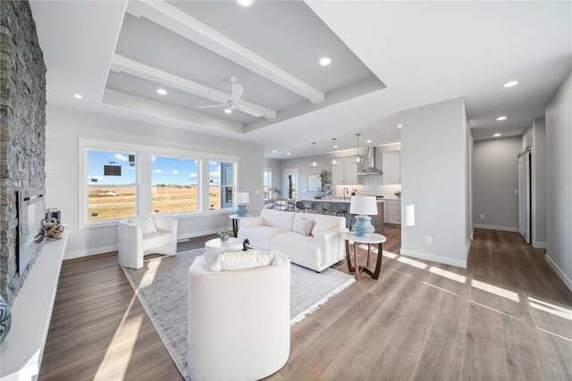 living room featuring ceiling fan, a tray ceiling, a stone fireplace, beamed ceiling, and light wood-type flooring