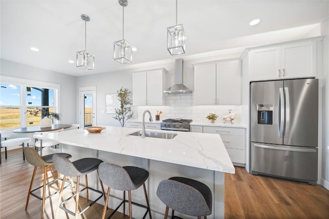 kitchen with sink, appliances with stainless steel finishes, an island with sink, wall chimney range hood, and white cabinets