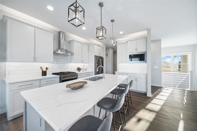 kitchen with wall chimney range hood, sink, appliances with stainless steel finishes, a center island with sink, and decorative light fixtures