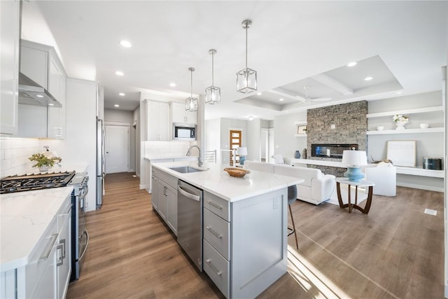 kitchen with appliances with stainless steel finishes, decorative light fixtures, sink, a center island with sink, and wall chimney exhaust hood