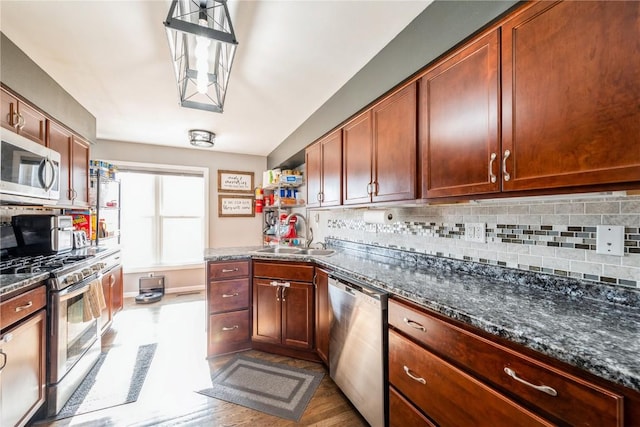 kitchen with appliances with stainless steel finishes, sink, backsplash, dark stone counters, and hanging light fixtures