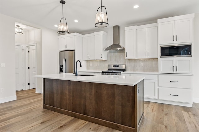 kitchen with sink, wall chimney range hood, appliances with stainless steel finishes, an island with sink, and decorative light fixtures