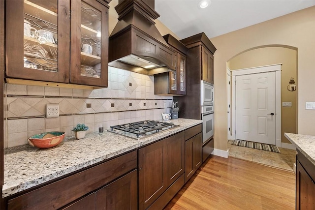 kitchen featuring stainless steel appliances, light hardwood / wood-style flooring, custom range hood, light stone counters, and decorative backsplash