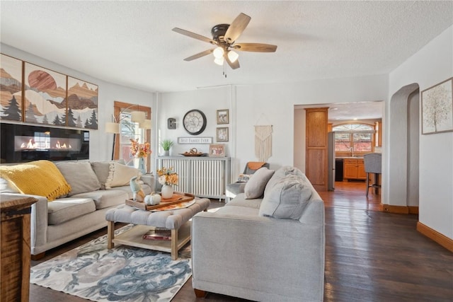 living area featuring baseboards, arched walkways, a glass covered fireplace, dark wood-style flooring, and a textured ceiling