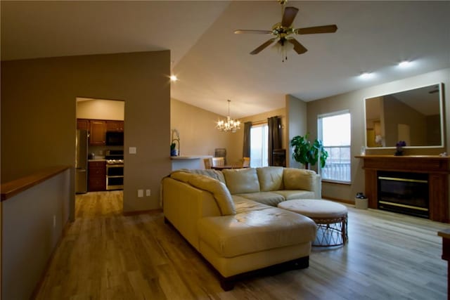 living room with vaulted ceiling, ceiling fan with notable chandelier, a glass covered fireplace, and light wood-style flooring