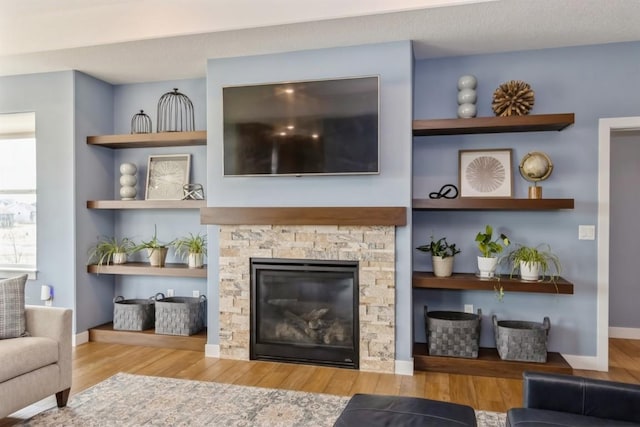 living room with built in shelves, a textured ceiling, a stone fireplace, wood finished floors, and baseboards