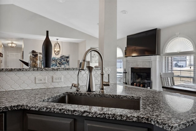 kitchen with a tiled fireplace, decorative backsplash, vaulted ceiling, a sink, and dark stone counters