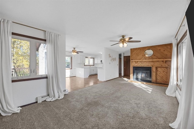 unfurnished living room with a ceiling fan, a glass covered fireplace, carpet floors, wooden walls, and a baseboard radiator