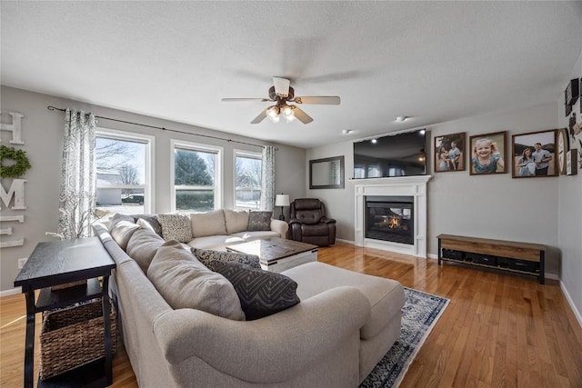 living room with ceiling fan, a textured ceiling, baseboards, light wood-type flooring, and a glass covered fireplace