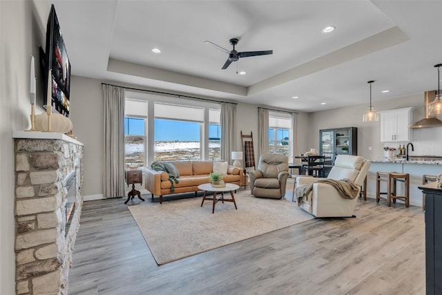 living room with ceiling fan, a stone fireplace, light wood-type flooring, and a raised ceiling