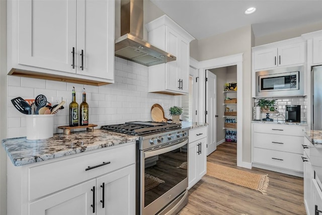 kitchen featuring light stone counters, light wood-style flooring, stainless steel appliances, white cabinetry, and wall chimney exhaust hood