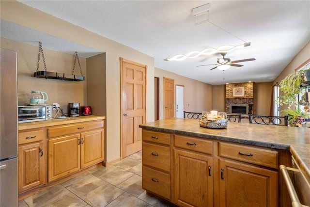 kitchen with a toaster, a fireplace, a ceiling fan, brown cabinetry, and open floor plan