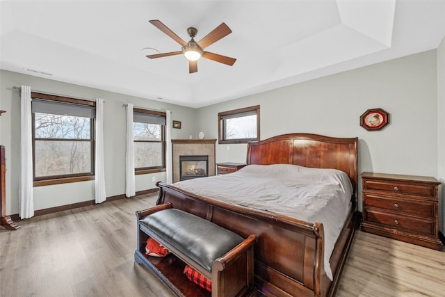 bedroom with baseboards, light wood-type flooring, a tray ceiling, a tile fireplace, and a ceiling fan