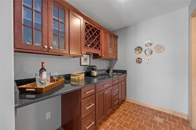 kitchen featuring brown cabinets, a sink, dark stone counters, glass insert cabinets, and baseboards