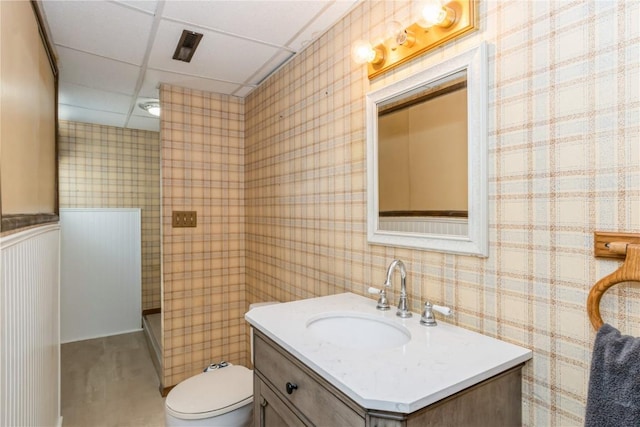 bathroom with a drop ceiling, toilet, wallpapered walls, and vanity