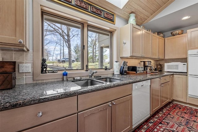 kitchen featuring tasteful backsplash, lofted ceiling with skylight, dark stone countertops, white appliances, and a sink