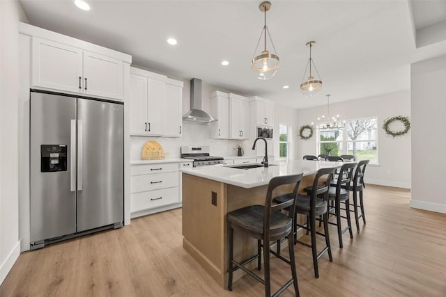 kitchen featuring a kitchen island with sink, a sink, white cabinets, appliances with stainless steel finishes, and wall chimney range hood