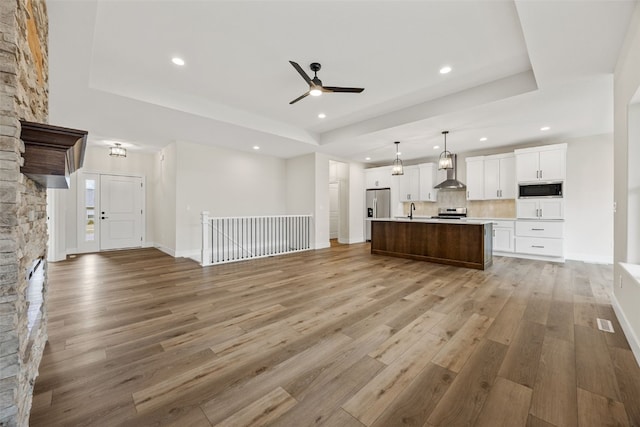 kitchen featuring light wood-type flooring, a tray ceiling, open floor plan, appliances with stainless steel finishes, and wall chimney exhaust hood