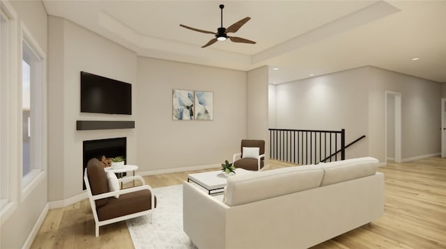 living area featuring a ceiling fan, baseboards, a fireplace, a raised ceiling, and light wood-type flooring