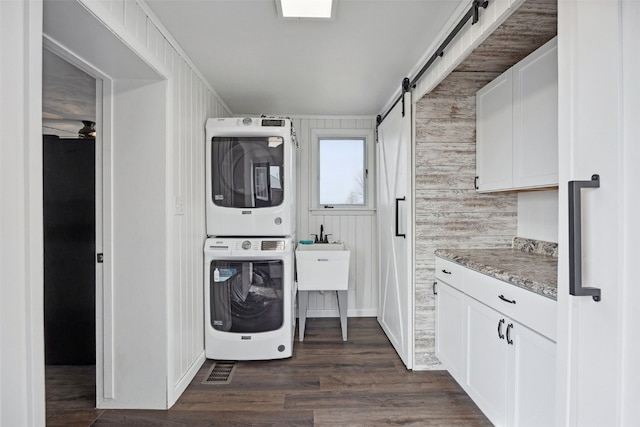 clothes washing area featuring dark wood-style floors, cabinet space, stacked washer and clothes dryer, and a barn door