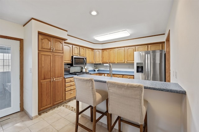 kitchen featuring brown cabinetry, light tile patterned flooring, ornamental molding, stainless steel appliances, and a kitchen breakfast bar