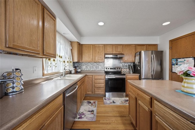 kitchen with a sink, under cabinet range hood, backsplash, appliances with stainless steel finishes, and light wood finished floors