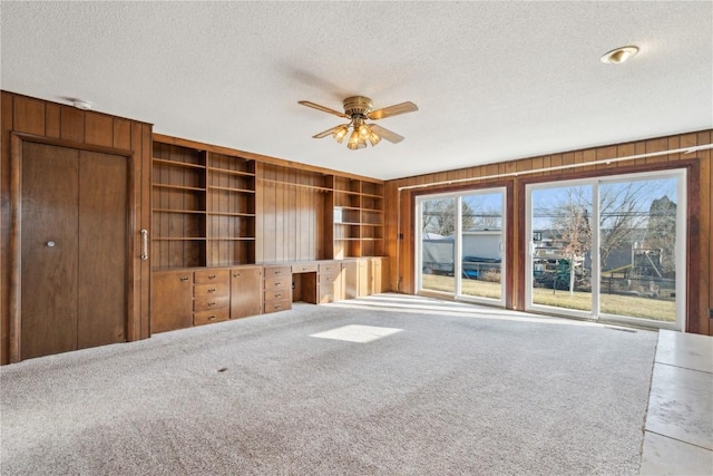 unfurnished living room featuring a textured ceiling, wood walls, carpet flooring, ceiling fan, and built in study area