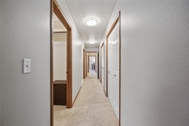 hall featuring a textured wall, light colored carpet, and baseboards