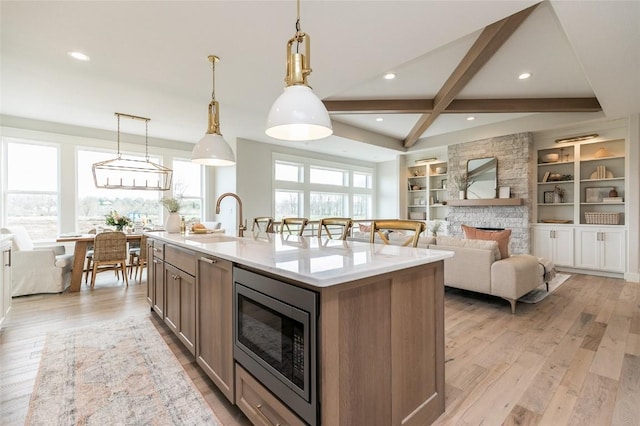 kitchen with light wood-type flooring, an island with sink, a sink, stainless steel microwave, and a fireplace