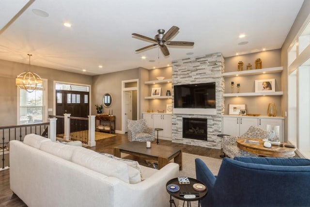 living room with dark wood finished floors, a stone fireplace, built in shelves, and ceiling fan with notable chandelier