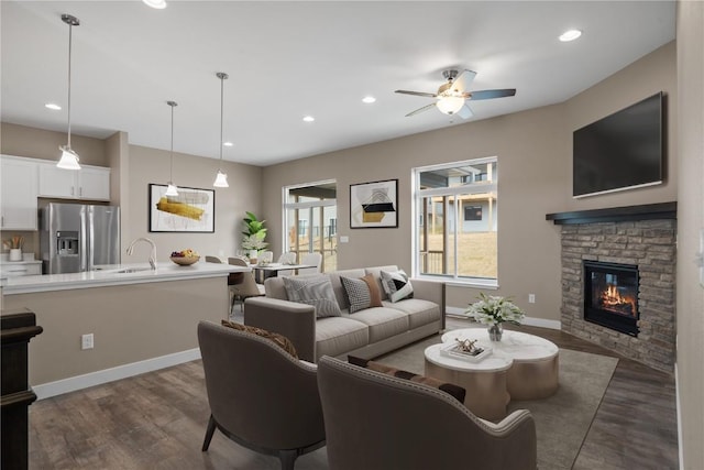 living area featuring baseboards, recessed lighting, dark wood-style flooring, ceiling fan, and a stone fireplace