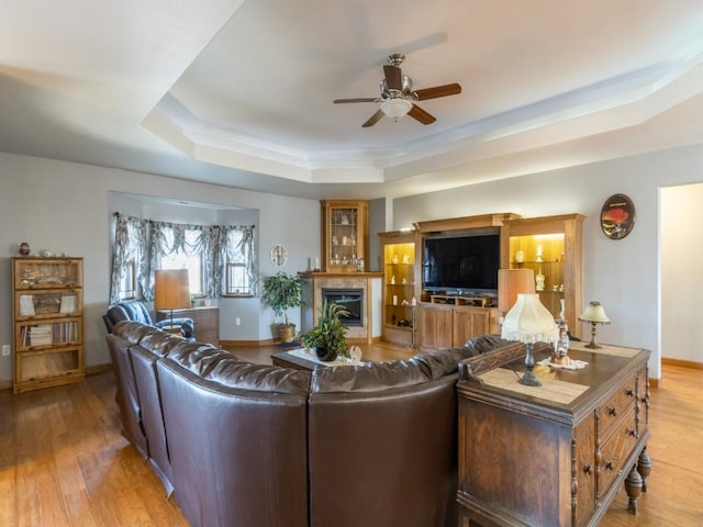 living area with baseboards, a tray ceiling, light wood-style flooring, a fireplace, and ceiling fan
