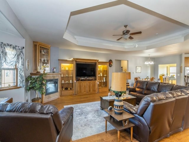 living area with a glass covered fireplace, a raised ceiling, ceiling fan with notable chandelier, and light wood finished floors