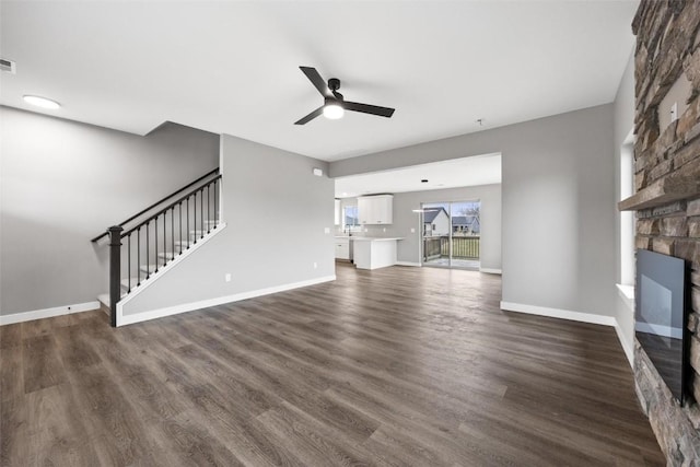 unfurnished living room featuring dark wood-type flooring, stairs, a ceiling fan, and a stone fireplace