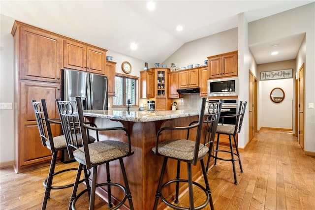 kitchen featuring appliances with stainless steel finishes, under cabinet range hood, brown cabinets, light stone counters, and light wood-style floors