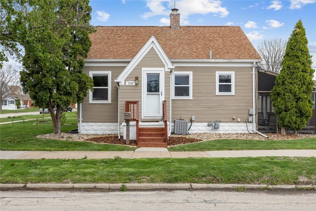 bungalow-style home with a shingled roof, a chimney, a front yard, and cooling unit