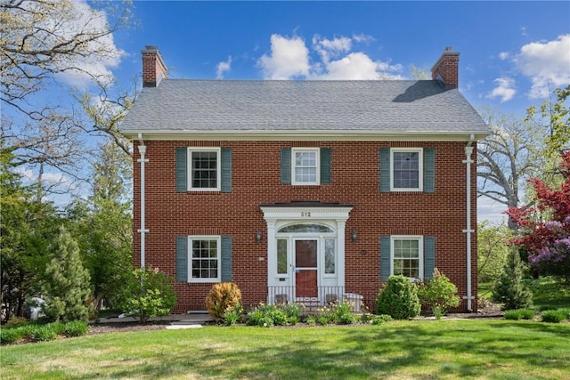 colonial inspired home featuring brick siding, roof with shingles, a front yard, and a chimney