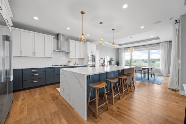 kitchen with wall chimney range hood, white cabinets, a tray ceiling, appliances with stainless steel finishes, and backsplash