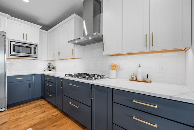 kitchen featuring wall chimney range hood, blue cabinetry, white cabinets, light wood-style floors, and stainless steel appliances