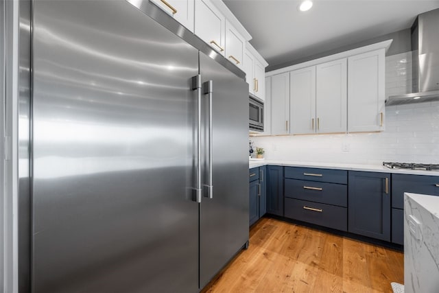 kitchen with white cabinets, wall chimney range hood, light wood finished floors, built in appliances, and blue cabinetry