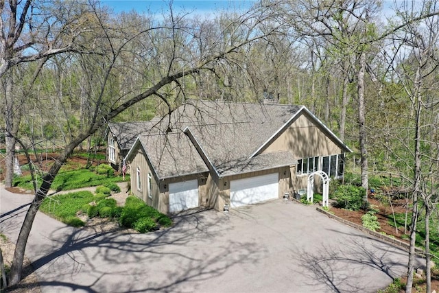 view of side of property with a garage, driveway, a wooded view, and a shingled roof