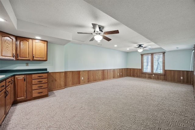 kitchen with a ceiling fan, light colored carpet, dark countertops, wainscoting, and brown cabinets